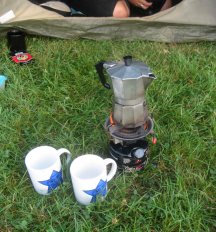 Kaffeemaschine auf einem Benzinkocher und zwei Tassen im Gras.